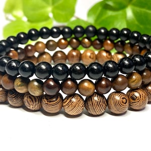 May include: Three stacked bracelets made of wooden beads. The top bracelet is black, the middle bracelet is brown and gold, and the bottom bracelet is brown with a striped pattern.