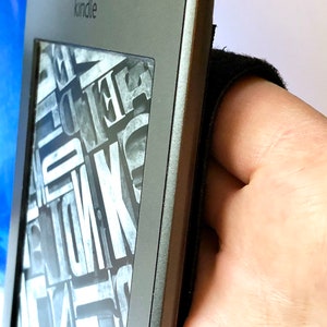 May include: A black and gray Kindle e-reader with the word "kindle" printed on the front. The screen is displaying a black and white pattern of letters.