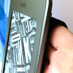 May include: A black Kindle e-reader with a textured gray cover and the word "kindle" printed on the front. The e-reader is held in a hand with a black strap.