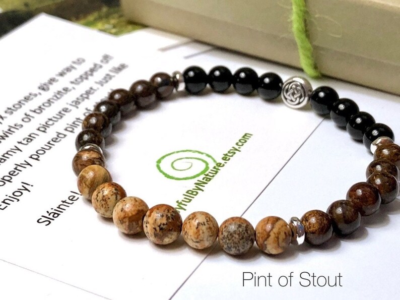 May include: A brown, black, and silver beaded bracelet with a Celtic knot charm. The bracelet is on a white card with the text "Pint of Stout" and a website address.