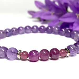 May include: A purple and pink amethyst gemstone bracelet with silver beads. The bracelet is made with round beads and has a gradient of color from light purple to dark purple.