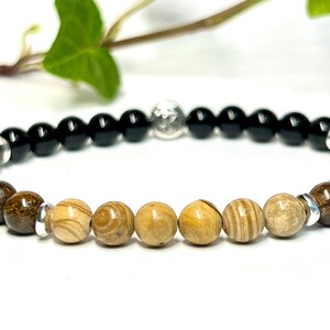 May include: A beaded bracelet with black, brown, and beige beads separated by silver spacers. The bracelet has a silver charm in the center.