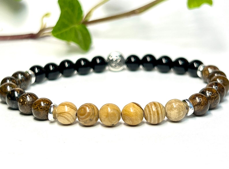 May include: A beaded bracelet with black, brown, and beige beads separated by silver spacers. The bracelet has a silver charm in the center.