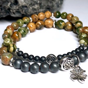 May include: A double-strand bracelet with green, brown, and black beads. The bracelet has a silver Celtic knot charm and a silver shamrock charm.