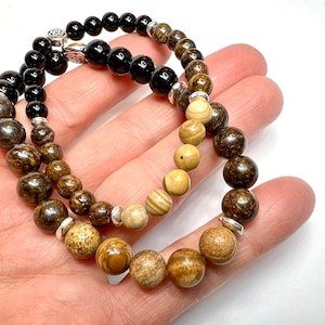 May include: Two beaded bracelets, one with black beads and the other with brown and beige beads. Both bracelets have silver accents.
