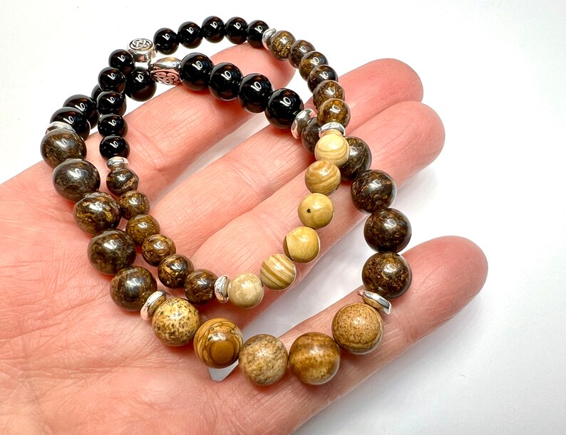 May include: Two beaded bracelets, one with black beads and the other with brown and beige beads. Both bracelets have silver accents.