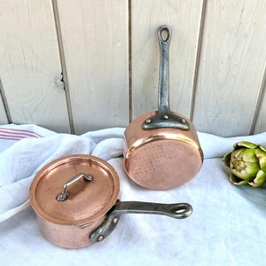 Peut inclure: Deux casseroles en cuivre avec des poignées gris métallisé. Une casserole est dotée d'un couvercle en cuivre assorti avec une petite poignée. Les casseroles sont posées sur un tissu blanc à rayures rouges, avec trois artichauts verts.