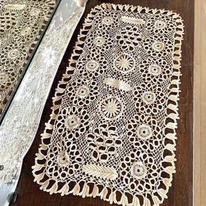 May include: A delicate, cream-colored crocheted table runner with a intricate pattern of circles and leaves. The runner is made of fine thread and has a delicate fringe around the edges.