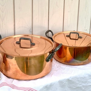 May include: Two copper pots with lids and black handles. The pots are on a white tablecloth with red stripes. There are onions and garlic cloves in front of the pots.