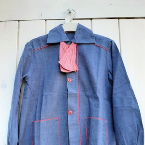 Puede incluir: Una camisa de algod&oacute;n azul con costuras rojas y una corbata de lazo a cuadros rojos y blancos. La camisa tiene dos bolsillos en la parte delantera y mangas largas.