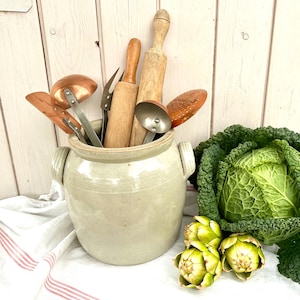 Op de afbeelding: Een beige keramieken pot met twee handvatten aan de zijkant bevat diverse keukengerei, waaronder twee houten deegrollers, koperen pollepels en een metalen vork. Naast de pot liggen een grote savooiekool en twee groene artisjokken.