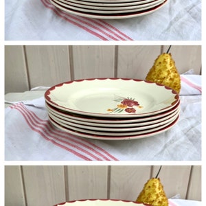 May include: A stack of six white ceramic plates with a red rim and a floral design. The plates are stacked on a white and red striped tablecloth.