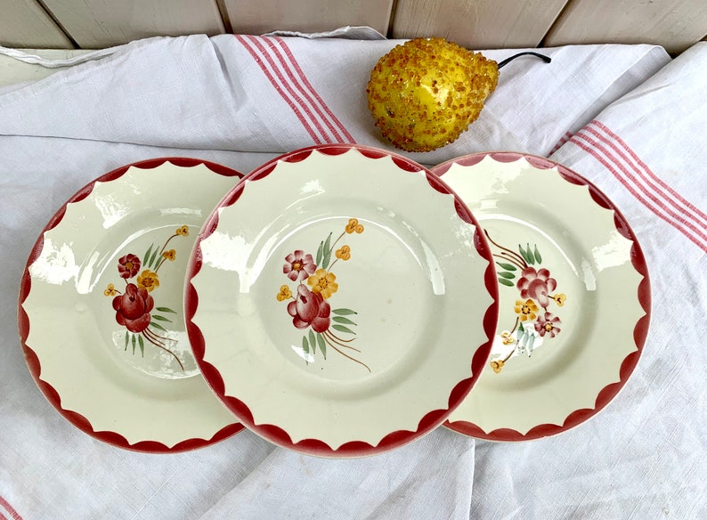 May include: Three white ceramic plates with scalloped edges and red trim. Each plate features a floral design with red, yellow, and green flowers.
