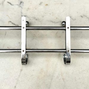 Puede incluir: Un perchero de pared horizontal hecho de metal cromado pulido. El perchero cuenta con dos barras horizontales paralelas conectadas por cuatro soportes verticales, cada uno terminado en un gancho curvo abajo y un peque&ntilde;o pomo esf&eacute;rico arriba.