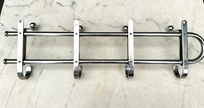 Puede incluir: Un perchero de pared horizontal hecho de metal cromado pulido. El perchero cuenta con dos barras horizontales paralelas conectadas por cuatro soportes verticales, cada uno terminado en un gancho curvo abajo y un peque&ntilde;o pomo esf&eacute;rico arriba.
