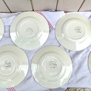 May include: Six white ceramic plates with a floral design and the words "Made in England" and "Anne" on the bottom. The plates are arranged in a circle on a white and pink striped tablecloth.