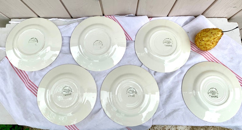 May include: Six white ceramic plates with a floral design and the words "Made in England" and "Anne" on the bottom. The plates are arranged in a circle on a white and pink striped tablecloth.