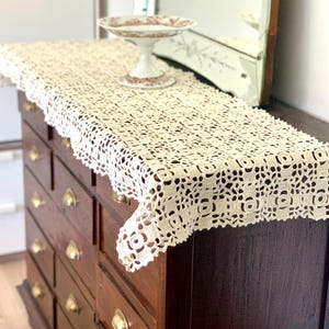May include: A white crocheted table runner with a delicate floral pattern, draped over a wooden dresser with multiple drawers and brass handles.