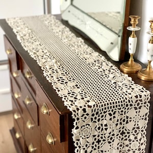 May include: Cream-colored, crocheted lace table runner with floral and geometric patterns. The runner is displayed on a dark wood dresser with brass hardware. A decorative mirror and candlesticks are in the background.