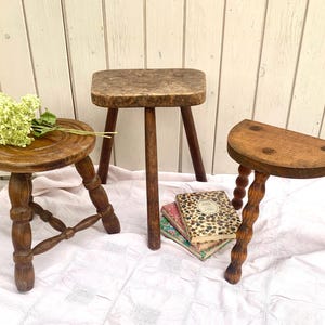 May include: Three antique wooden stools of varying designs. One stool is round with turned legs, another has a square top, and the third has a half-moon shape with spiral legs. A small bouquet of flowers rests on one stool.