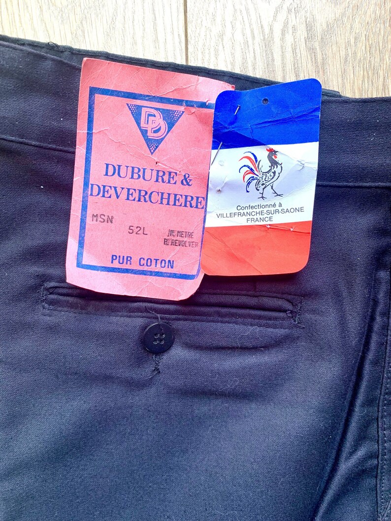 May include: A red and white label with the text "DUBURE & DEVERCHERE" and "MSN 52L" in black. The label also includes a red triangle with a black letter "D" inside. A smaller blue, white, and red label with a rooster illustration and the text "Confectionn&eacute; &agrave; VILLEFRANCHE-SUR-SAONE FRANCE" is attached to the larger label.