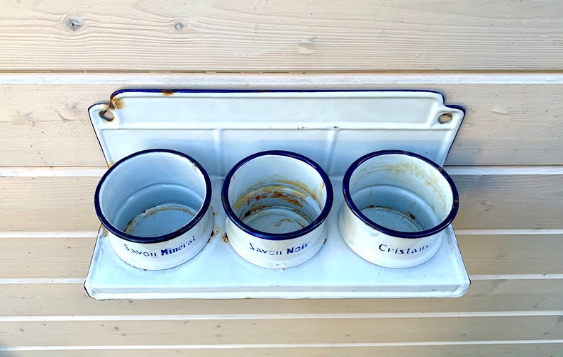 May include: White enamel wall-mounted shelf with three round white enamel containers. The containers have blue trim and are labeled "Savon Mineral", "Savon Noir", and "Cristaux".