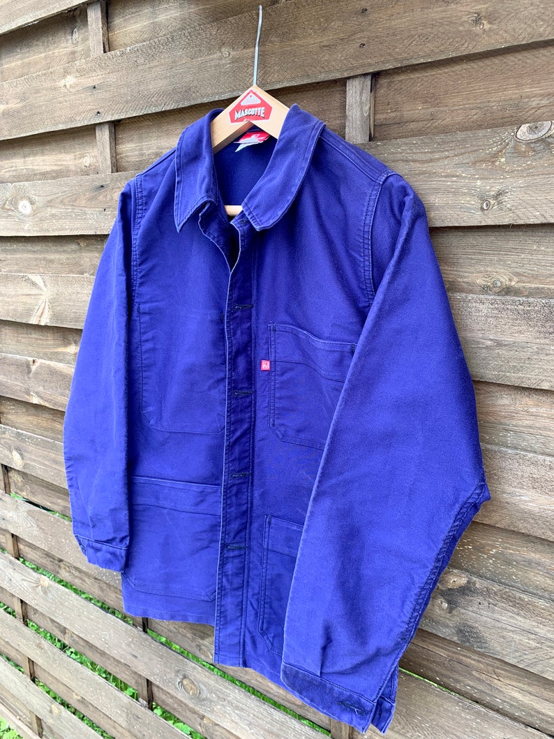 Pode incluir: Uma jaqueta de trabalho azul com fecho de bot&otilde;es e um bolso no peito. A jaqueta tem uma etiqueta que diz "Marquette" no cabide.