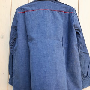 Puede incluir: Una camisa de manga larga azul oscuro con un ribete rojo alrededor del cuello y por el delantero. La camisa est&aacute; colgada en una percha de madera.
