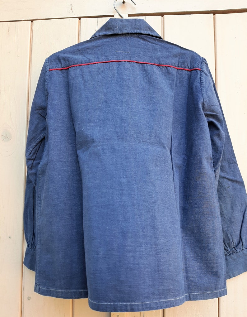 Puede incluir: Una camisa de manga larga azul oscuro con un ribete rojo alrededor del cuello y por el delantero. La camisa est&aacute; colgada en una percha de madera.