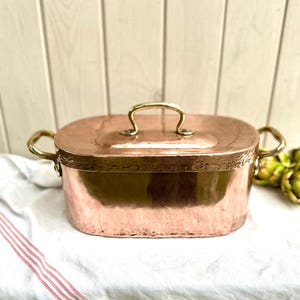 Puede incluir: Una cacerola ovalada de cobre con tapa y asas de latón. El plato está sobre un paño blanco con rayas rojas. El cobre tiene un acabado cálido y reflectante. La tapa tiene un asa de latón.