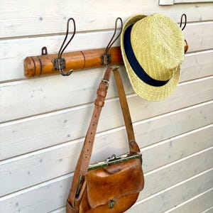 May include: A wooden coat rack with three hooks, a straw hat hanging on one hook, and a brown leather bag hanging on another hook.