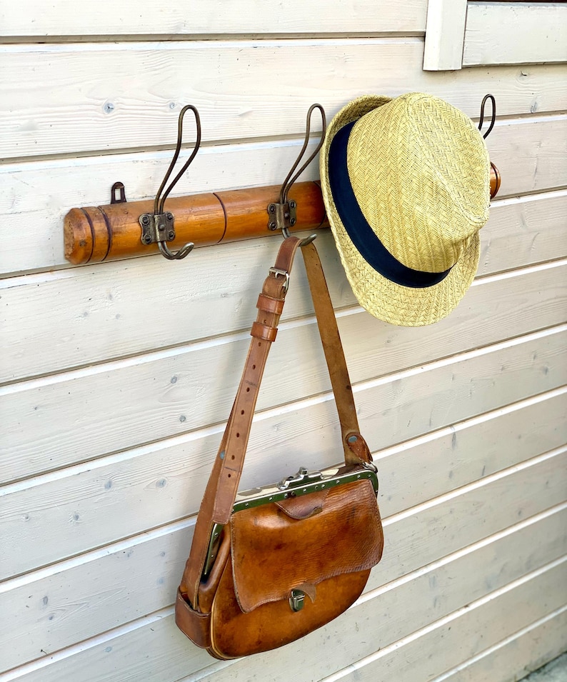 May include: A wooden coat rack with three hooks, a straw hat hanging on one hook, and a brown leather bag hanging on another hook.
