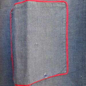 Puede incluir: Primer plano de una tela vaquera azul con un bolsillo cosido rojo. El bolsillo es de forma cuadrada y tiene un borde cosido rojo.
