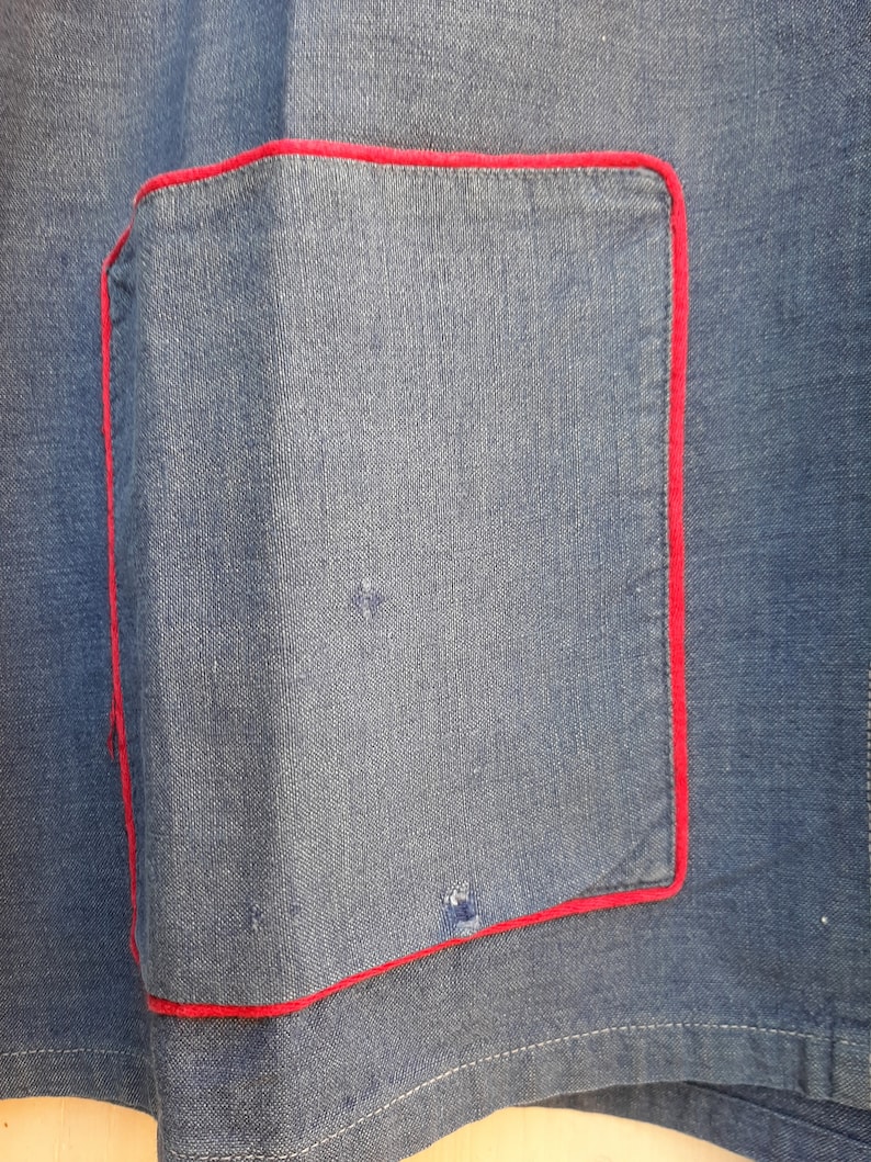 Puede incluir: Primer plano de una tela vaquera azul con un bolsillo cosido rojo. El bolsillo es de forma cuadrada y tiene un borde cosido rojo.