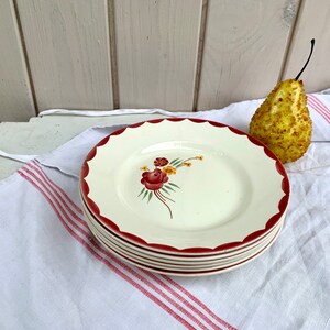 May include: A stack of four white ceramic plates with a red rim and a floral design. The plates have a scalloped edge and are stacked on a white and red striped tablecloth.