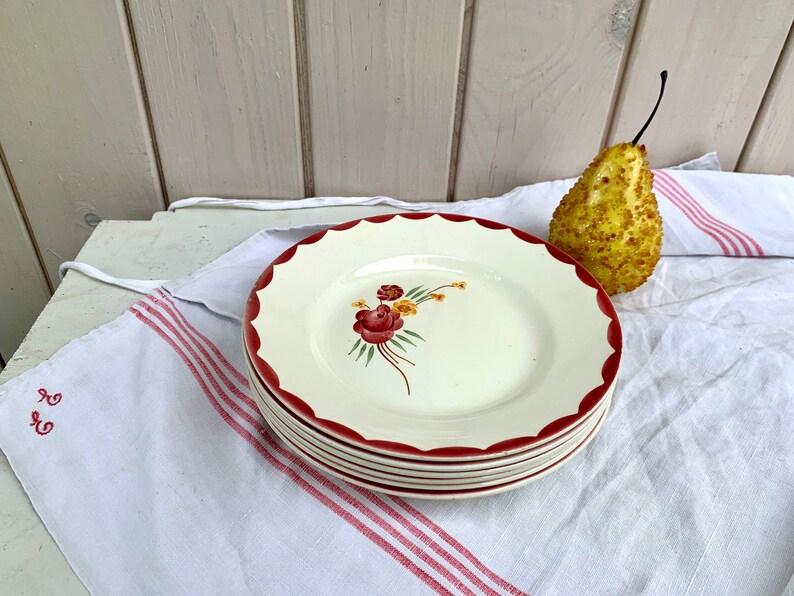 May include: A stack of four white ceramic plates with a red rim and a floral design. The plates have a scalloped edge and are stacked on a white and red striped tablecloth.
