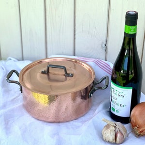 May include: A copper pot with a lid and two handles sits on a white and red striped tea towel. A bottle of Côtes du Rhône wine, a whole onion, and a head of garlic are also on the towel.
