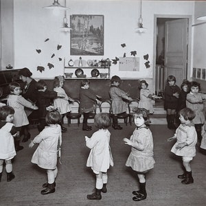 Puede incluir: Una fotograf&iacute;a en blanco y negro de un aula de ni&ntilde;os en c&iacute;rculo, tom&aacute;ndose de la mano y cantando. Los ni&ntilde;os llevan vestidos y camisas. El aula tiene una pizarra, una estanter&iacute;a con platos y una imagen en la pared.