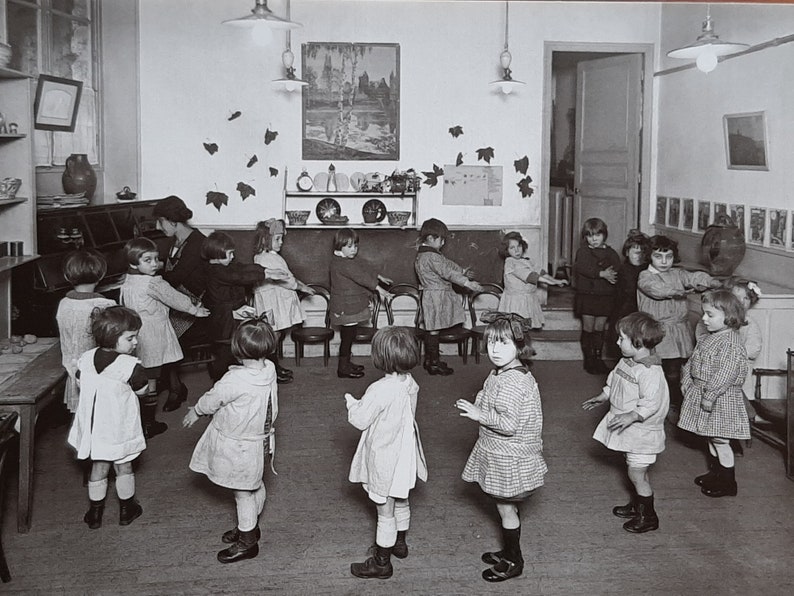 Puede incluir: Una fotograf&iacute;a en blanco y negro de un aula de ni&ntilde;os en c&iacute;rculo, tom&aacute;ndose de la mano y cantando. Los ni&ntilde;os llevan vestidos y camisas. El aula tiene una pizarra, una estanter&iacute;a con platos y una imagen en la pared.