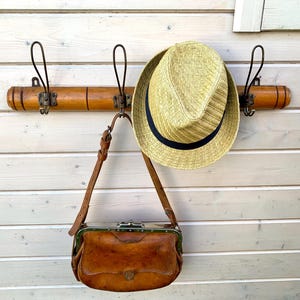 May include: A wooden coat rack with three hooks and a brown leather purse hanging from one of the hooks. A straw hat is hanging from another hook.