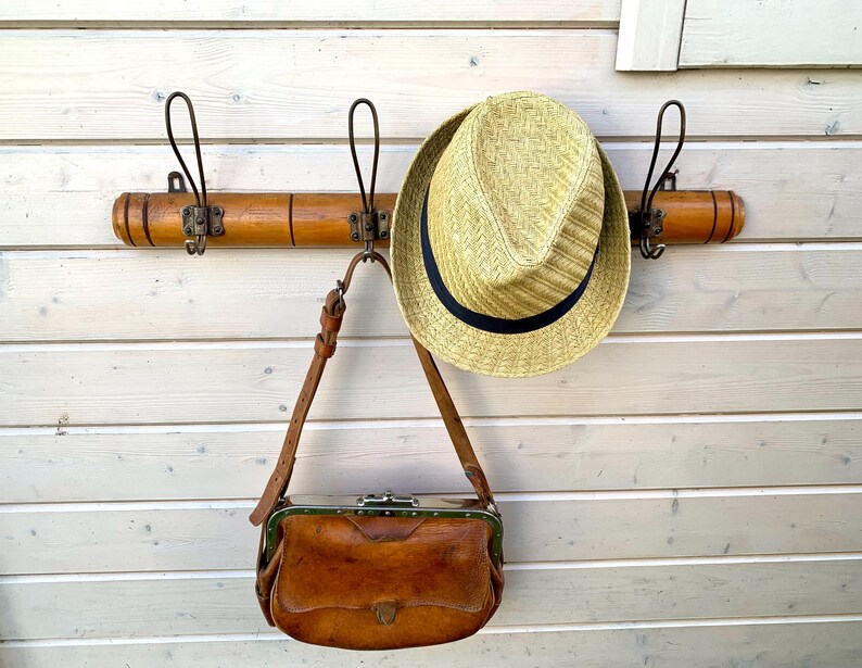 May include: A wooden coat rack with three hooks and a brown leather purse hanging from one of the hooks. A straw hat is hanging from another hook.