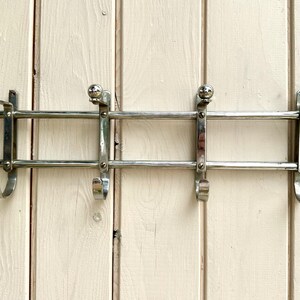 Puede incluir: Un perchero de pared hecho de metal cromado pulido. El perchero cuenta con dos barras horizontales conectadas por cuatro soportes verticales, cada uno terminado en un gancho con punta de bola arriba y un gancho curvo abajo para colgar.