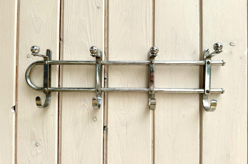 Puede incluir: Un perchero de pared hecho de metal cromado pulido. El perchero cuenta con dos barras horizontales conectadas por cuatro soportes verticales, cada uno terminado en un gancho con punta de bola arriba y un gancho curvo abajo para colgar.