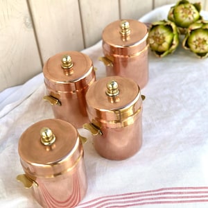 Könnte beinhalten: Vier kupferfarbene Dosen mit Deckeln und kleinen Griffen. Jede Dose hat einen goldfarbenen Knopf auf dem Deckel. Die Dosen sind auf einem weißen Tuch mit roten Streifen angeordnet.