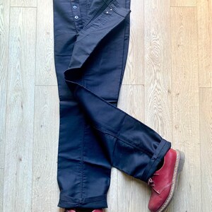 May include: A pair of dark blue jeans with a button fly and a cuffed hem. The jeans are folded and laid out on a wooden floor. Two red leather shoes are placed on the floor, one on each leg of the jeans.