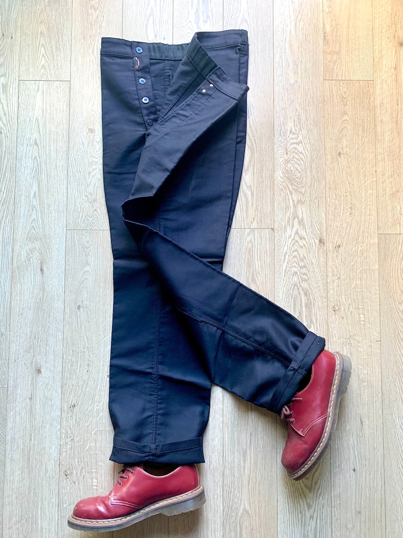 May include: A pair of dark blue jeans with a button fly and a cuffed hem. The jeans are folded and laid out on a wooden floor. Two red leather shoes are placed on the floor, one on each leg of the jeans.