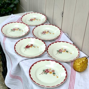 May include: Set of six white ceramic plates with a scalloped edge and a floral design in shades of red, yellow, and green. The plates are arranged on a white tablecloth with red stripes.