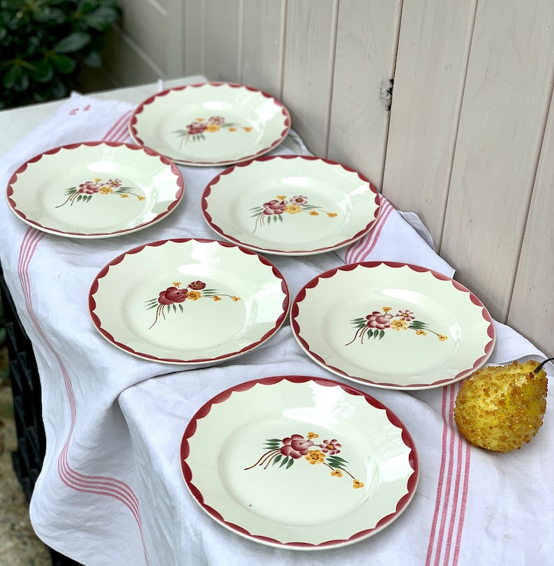 May include: Set of six white ceramic plates with a scalloped edge and a floral design in shades of red, yellow, and green. The plates are arranged on a white tablecloth with red stripes.