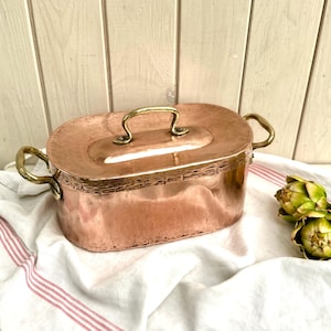 May include: A copper oval cooking pot with a lid and brass handles. The pot sits on a white cloth with red stripes, next to three green artichokes. The pot has a shiny, reflective surface.