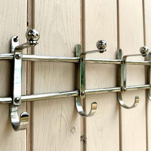 Puede incluir: Un perchero de pared fabricado en metal con acabado cromado pulido. El soporte presenta una estructura horizontal de doble barra con cuatro ganchos orientados hacia abajo y cuatro perchas orientadas hacia arriba, cada una rematada con un peque&ntilde;o remate esf&eacute;rico.